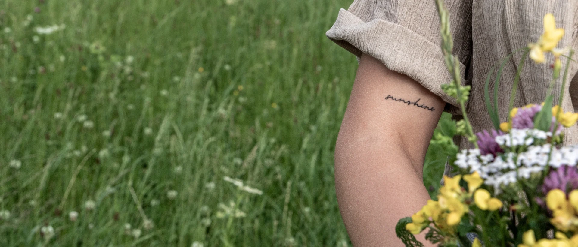 Arm mit Sunshine-Tattoo und einem Blumenstrauß auf einer grünen Wiese