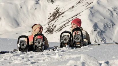 Zwei Personen mit Schneeschuhen sitzen lachend im Schnee vor Bergen