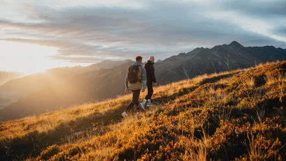 Zwei Wanderer steigen bei Sonnenuntergang einen grasbewachsenen Hügel mit Bergen im Hintergrund hinauf