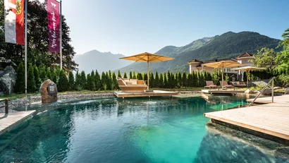 Ein Sommertag am türkisfarbener Naturpool mit Relaxmöbel und einem aufgespannten Sonnenschirm im Hintergrund mit Blick auf die umliegenden Berge