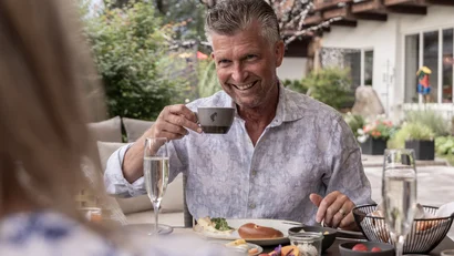 Ein Paar sitzt am Frühstückstisch auf der Sonnenterrasse im Hotel Gassner, der Mann hält eine Kaffeetasse in der Hand und lächelt seiner Frau entgegen.