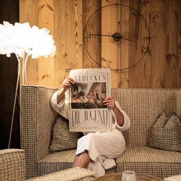 Person in bathrobe reading newspaper on sofa in front of wooden wall with large clock
